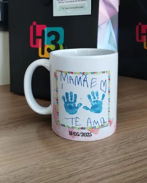 Caneca branca com o desenho de uma criança: duas mãozinhas azuis, a frase Mamãe te amo e a data 11/05/2025, na frente da sacola preta da H3
