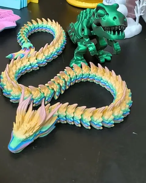 Dois dragões articulados em tons pastel e um dinossauro verde, impressos em 3D, sobre a bancada
